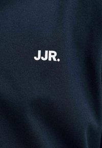 Λευκό κείμενο "JJR." τυπωμένο σε σκούρο μπλε υφασμάτινο φόντο με υφή, πιθανόν ρούχο.
