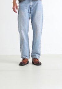 Brūni ādas zābaki, gaiši zili taisni džinsi, viegli nodiluši. Fons ir neitrāls, izceļot apavus un apģērbu.