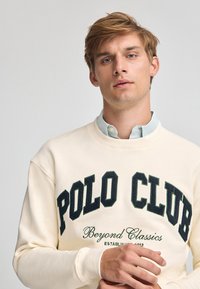 Felpa crema con la scritta "POLO CLUB" in testo verde scuro, con "Beyond Classics" ricamato sotto. Indossata sopra una camicia a righe con colletto.