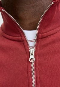 Felpa rossa con zip sul davanti, caratterizzata da una cerniera argentata, colletto a costine e un tessuto testurizzato. Sotto, c'è una maglietta bianca con righe rosse.