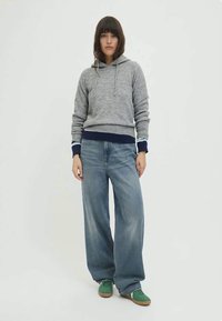 Grauer Kapuzenpulli mit einer Fronttasche und marineblauem Saum, kombiniert mit weit geschnittenen blauen Jeans. Akzentgrüne Sneakers runden das Outfit ab.