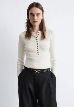 Geripptes cremefarbenes Langarmoberteil mit Knopfleiste, kombiniert mit hochtaillierter schwarzer Hose und einem schwarzen Gürtel mit goldener Schnalle.