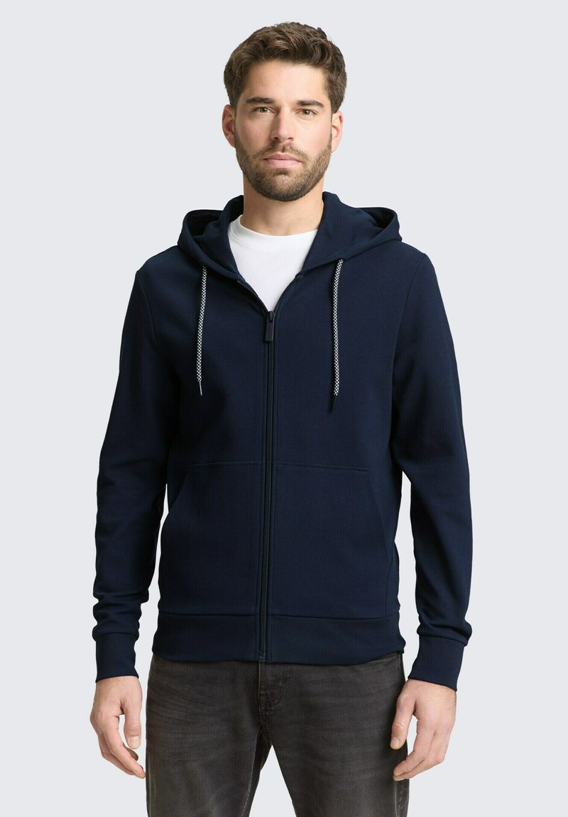 Uomo che indossa una felpa con cappuccio blu navy con cerniera e lacci bianchi sopra una maglietta bianca, in piedi davanti a uno sfondo grigio chiaro uniforme.