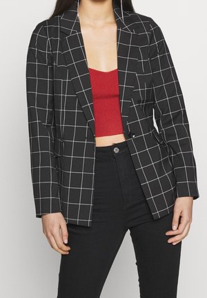 Mujer con blazer de cuadros en blanco y negro sobre top corto rojo de canalé y pantalones negros de talle alto, de pie con las manos relajadas.