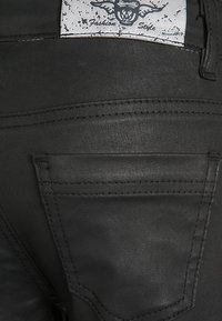 Pantalones de mezclilla negros con una superficie texturizada, costuras prominentes y un único bolsillo trasero con una etiqueta que muestra "Estilo de Moda."