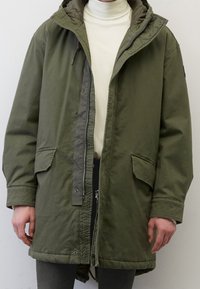 Olivgrön parka-jacka med dragkedja och tryckknappar, stora framfickor, en vadderad innerfoder och en huva med justerbara snoddar.