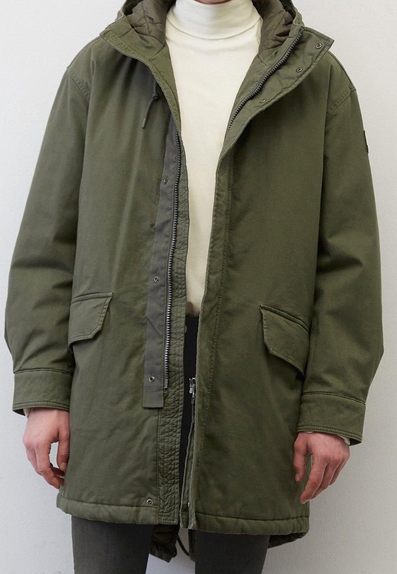Olivgrön parka-jacka med dragkedja och tryckknappar, stora framfickor, en vadderad innerfoder och en huva med justerbara snoddar.