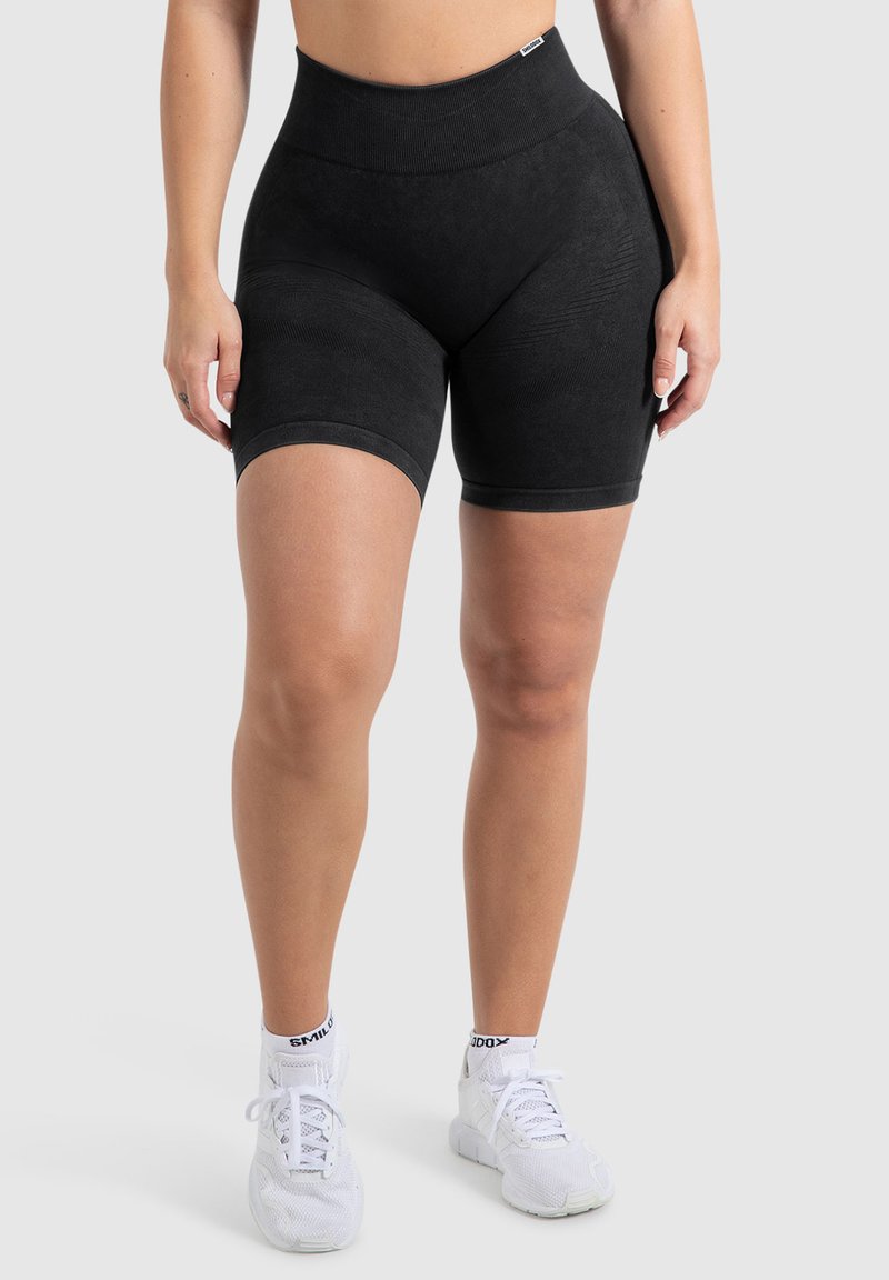 Zwarte high-waisted shorts met een zachte textuur, geribbelde tailleband en contrasterende details aan de zijkanten. Gecombineerd met witte sportschoenen.