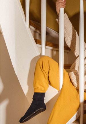 Kind met gele broek en zwarte sokken zit op de trap en houdt met de hand en het been tussen de spijlen een wit traphekje vast.