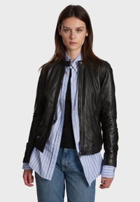 Crna kožna motoristička jakna s okruglim ovratnikom, patentnim zatvaračem sprijeda i uskim rukavima, složena preko svijetloplavog prugastog košulje i tamnog majice bez rukava.