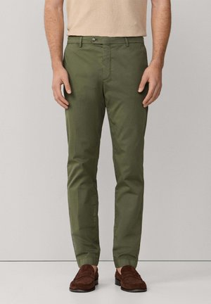 Hombre con pantalones rectos verde oliva y mocasines de ante marrón oscuro, de pie contra un fondo liso de color gris claro.