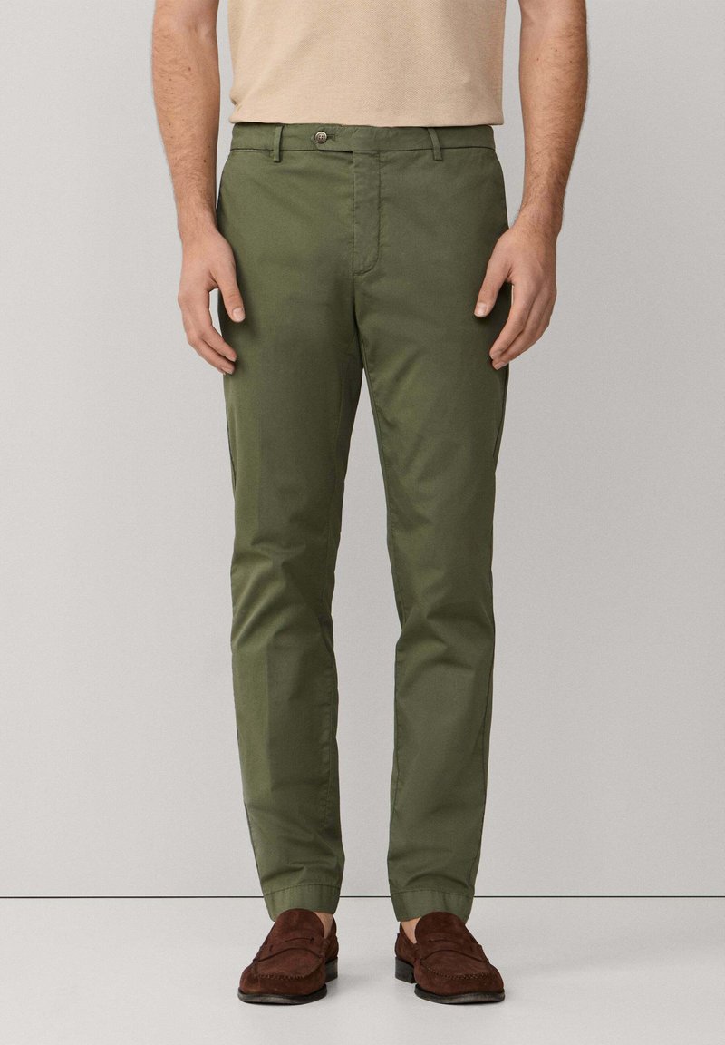 Muškarac obučen u ravne hlače maslinasto zelene boje i tamnoplave kožne mokasine, stoji na jednostavnoj svijetlosivoj pozadini.