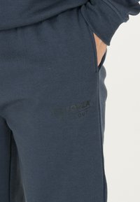 Mörkblå sweatpants tillverkade av mjukt material, med en svart logotyp tryckt på sidan och sidofickor för extra funktionalitet.