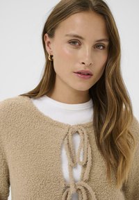 Smėlio spalvos tekstūruotas megztinis su apvalia iškirpte ir raišteliu, dėvimas ant baltos ribuotos palaidinės. Papildytas natūralių plaukų ir subtilių papuošalų akcentais.