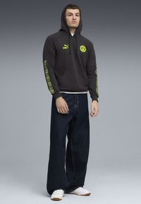 Ung mand iført sort hættepullover med gule logoer, brede mørke jeans og hvide sneakers, der står mod en grå baggrund.
