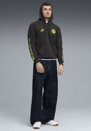 Ung mand iført sort hættepullover med gule logoer, brede mørke jeans og hvide sneakers, der står mod en grå baggrund.