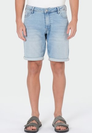 Mann trägt hellblaue hochgekrempelte Jeansshorts, ein weißes Hemd und graue Sandalen und steht vor einem schlichten weißen Hintergrund.
