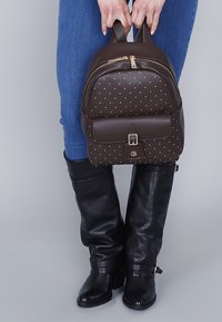 Mochila de cuero marrón con tachuelas doradas, cierre de cremallera y un bolsillo con solapa en la parte frontal. Combinada con jeans azules y botas negras hasta la rodilla.