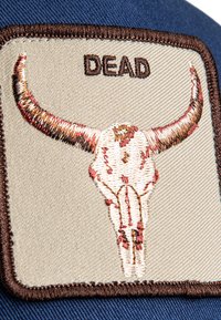 Patch ricamato con un teschio di toro con corna su uno sfondo beige. La parola "DEAD" è cucita sopra il teschio con filo marrone.