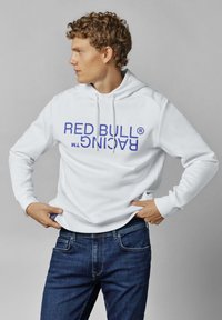 Sudadera blanca con un gran logo azul de "RED BULL RACING". Tanto la capucha como las mangas son largas, con cordones. Combinada con jeans azul oscuro.