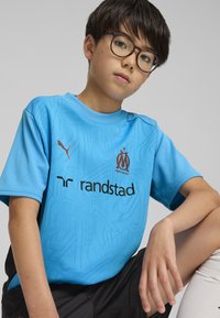 Gaiši zils krekls ar reljefa rakstiem, kas rotāts ar Puma logo un Marseļas emblēmu. Melni šorti un baltas zeķes papildina apģērbu.