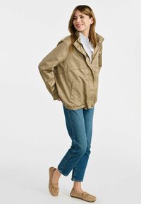 Khaki lekka kurtka z wysokim kołnierzem, zatrzaskami i dwiema przednimi kieszeniami, połączona z niebieskimi dżinsami i beżowymi butami wsuwanymi.