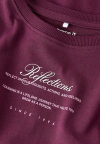 Bordowa bawełniana bluza z okrągłym dekoltem. Posiada biały napis: "Refleksje... rozwijaj się jako osoba. OD 1996" w różnych czcionkach.