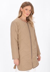 Giacca trapuntata beige con zip frontale, maniche lunghe e tasche laterali. Tessuto liscio con motivo di cuciture diagonali.