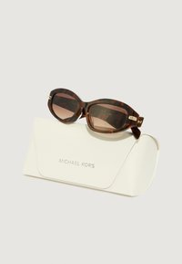 Occhiali da sole tartarugati con montatura spessa, lenti marroni e dettagli in oro, appoggiati su una custodia bianca con il logo "Michael Kors" in rilievo.
