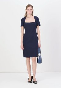 Temno modra obleka z oprijetim krojem, kratkimi rokavi in kvadratnim izrezom. Gladka tekstura z dolžino do kolen, v kombinaciji z majhno modro torbico.