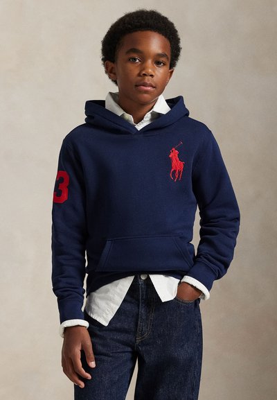 Junge trägt dunkelblauen Hoodie mit rotem Logo und Nummer 3, weißes Hemd darunter und dunkelblaue Jeans, steht mit einer Hand in der Tasche.