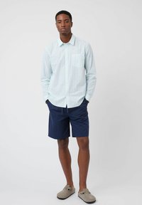 Camisa de rayas azul claro de manga larga con un bolsillo en el pecho, combinada con pantalones cortos azul marino y zapatos de lona grises. Tela suave, ajuste casual.