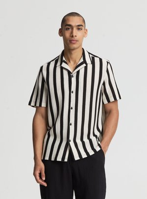 Giovane uomo con indosso una camicia a maniche corte a righe verticali bianche e nere e pantaloni neri, in piedi davanti a uno sfondo chiaro e semplice.