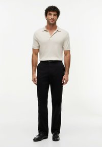 Uomo in piedi con polo beige a maniche corte, pantaloni neri, cintura nera e scarpe nere su uno sfondo bianco semplice.