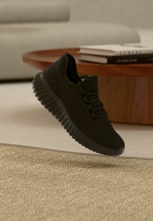 Adidaș sport negru cu talpă texturată, suspendat deasupra unui covor bej țesut, cu o masă din lemn și cărți stivuite în fundal.