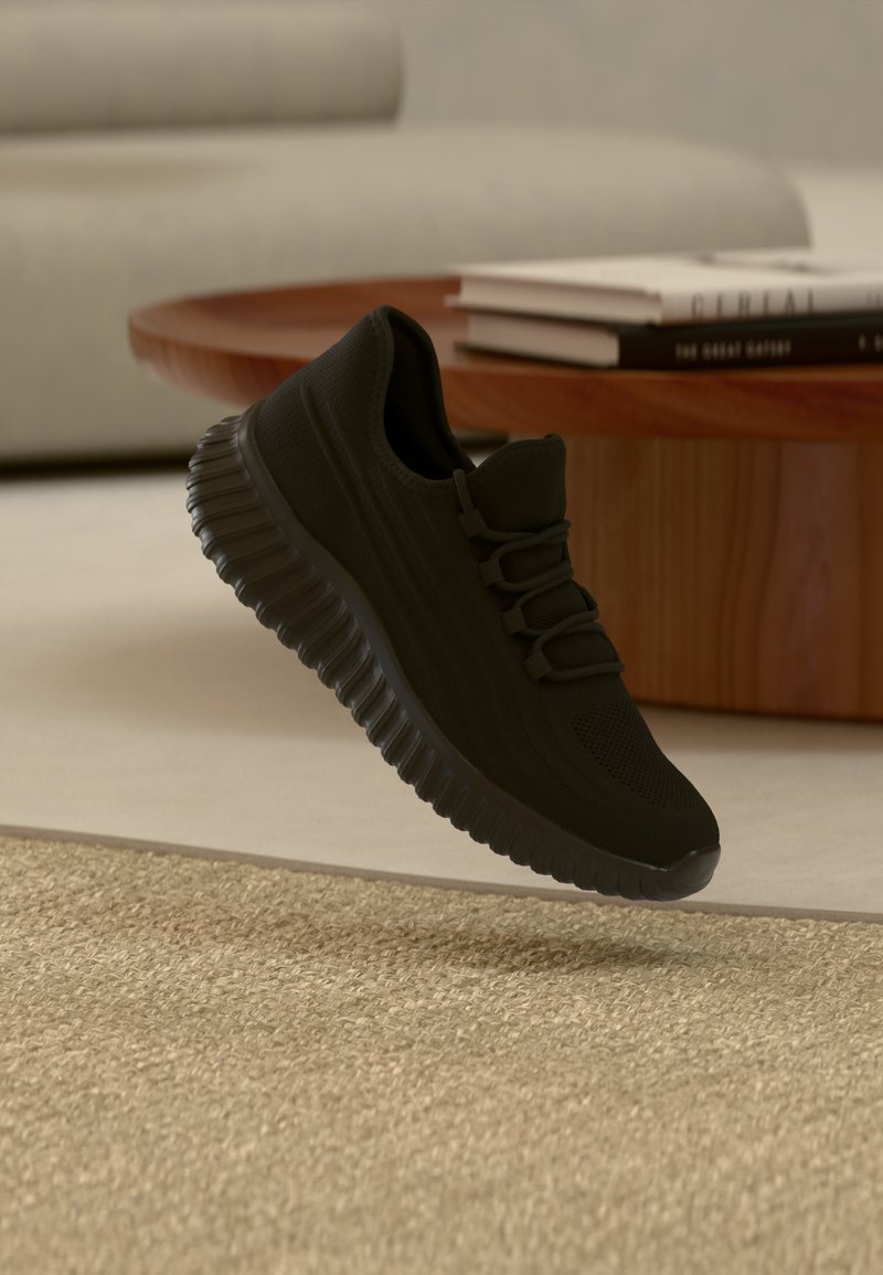 Baskets athlétiques noires avec semelle texturée suspendues au-dessus d'un tapis en lin beige tissé, avec une table en bois et des livres empilés en arrière-plan.