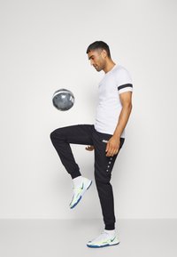 Witte sportshirt met zwarte mouwaccenten, zwarte joggerbroek met knopen en helder witte en blauwe sportschoenen; een grijze voetbal is op één voet in balans.