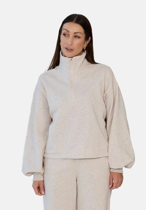 Kobieta ubrana w jasnobeżową bluzę zapinaną na zamek z wysokim kołnierzem oraz pasujące spodnie, patrzy lekko w lewo na jednolitym tle.