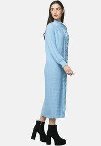 Lyseblå, lang, strikket kjole med høj halsudskæring og tekstureret fletmønster; kombineret med sorte, chunky ankelstøvler med hæl.