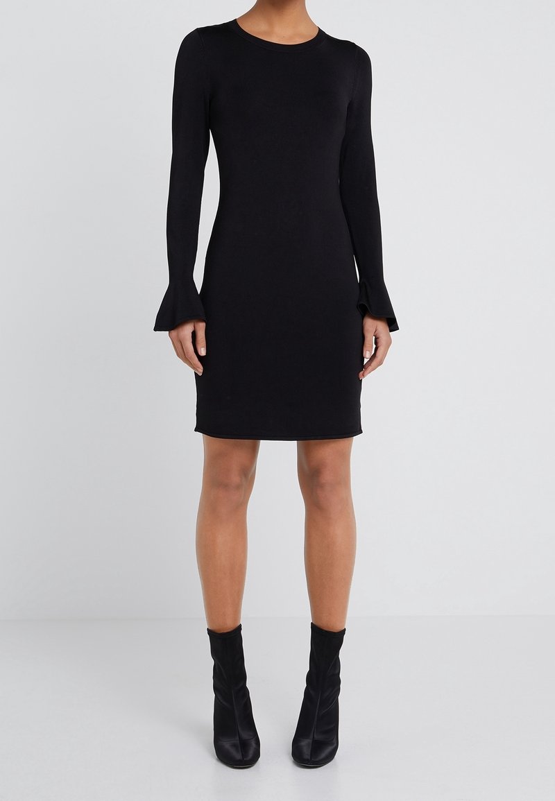 Femme portant une robe noire cintrée à manches longues avec des poignets évasés et des bottines noires, debout sur un fond uni.