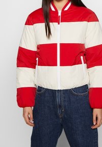 Giacca bomber a righe rosse e crema con zip frontale, polsini elasticizzati e tasche laterali. Tessuto leggero e trapuntato con un design sportivo.