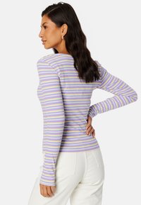 Długi rękaw, dopasowana bluzka w lawendowe i białe paski, wykonana z miękkiego materiału. Posiada okrągły dekolt i ribbową strukturę. Widoczne białe spodnie.