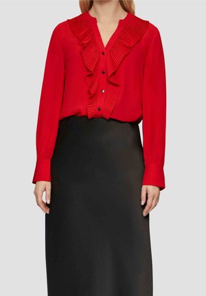 Vrouw draagt een rode blouse met lange mouwen en ruches aan de voorkant en een zwarte hoge taille rok tegen een effen lichte achtergrond.