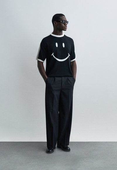 Άνδρας που φοράει μαύρο παντελόνι με φαρδιά πόδια, μαύρο πουλόβερ με κοντά μανίκια και λευκό πρόσωπο με χαμόγελο, μαύρα γυαλιά ηλίου και μαύρα παπούτσια, στέκεται με τα χέρια στις τσέπες.