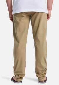 Beige, lightweight trousers with a straight cut; visible stitch lines; worn with black flip-flops; plain back design without pockets.