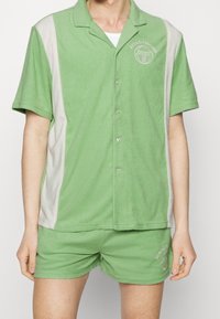 Conjunto de dos piezas verde hecho de tela suave; camisa de cuello corto con paneles laterales gris claro y pantalones cortos a juego, que destacan por los acentos del logo.