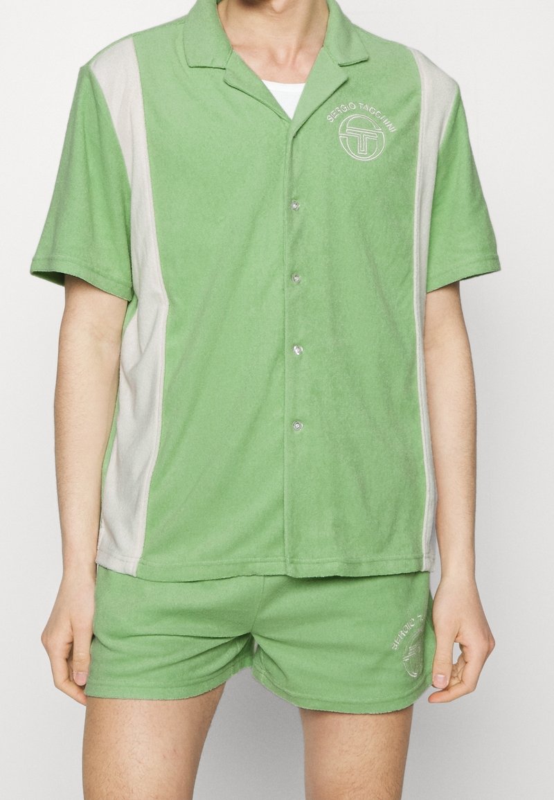 Conjunto de dos piezas verde hecho de tela suave; camisa de cuello corto con paneles laterales gris claro y pantalones cortos a juego, que destacan por los acentos del logo.