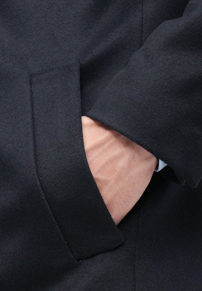 Cappotto in lana nera con finitura strutturata, dotato di una tasca laterale con una mano appoggiata casualmente al suo interno. Design semplice e pulito.