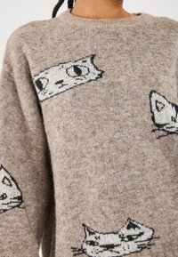 Pull beige en tricot avec motifs de chats noirs, col rond, tissu doux et texturé. Le design comprend diverses expressions et formes de chats.