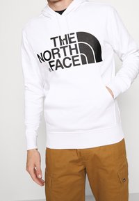 Persona che indossa una felpa bianca con un grande logo nero "The North Face" e pantaloni cargo marroni, con una mano in tasca della felpa.